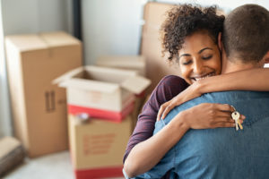 Couple hugging, happy and in love as they move into their new home after marriage counseling in Fairfax, VA. There are moving boxes all over the background and one partner has keys in her hand. Couples therapy in Fairfax, VA and Woodbridge, VA can help you reconnect and feel more connected in your marriage.
